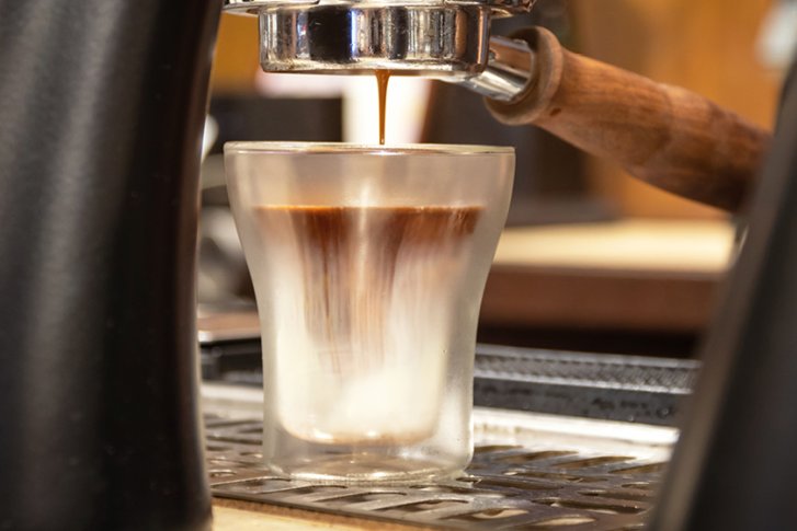 Espressomaschine füllt Glas mit Kaffee, umgeben von metallischen Teilen und unscharfem Hintergrund.