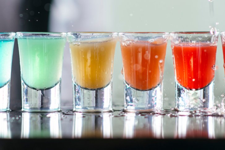 Acht bunte Shots stehen in einer Reihe auf einem nassen Tisch.