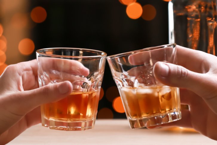 Zwei Hände stoßen Whiskygläser an, im Hintergrund leuchten orange Lichter unscharf.