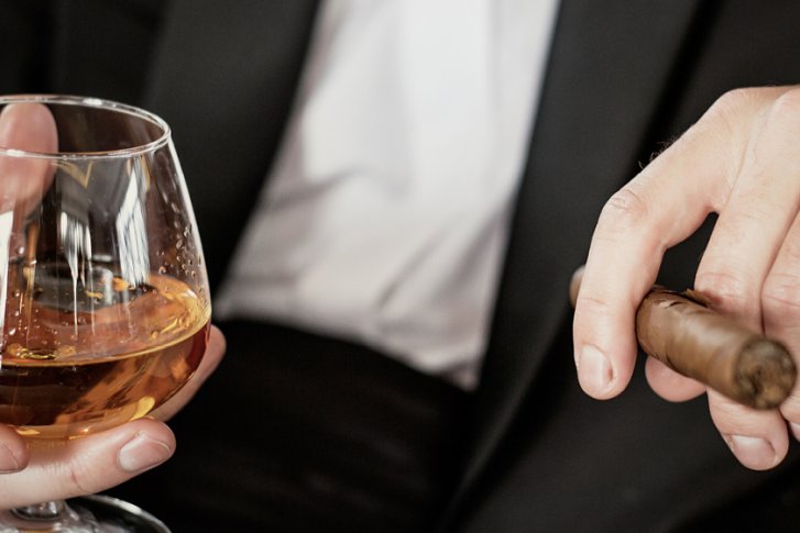 Ein Mann hält ein Cognacglas und eine Zigarre, in eleganter Abendgarderobe.