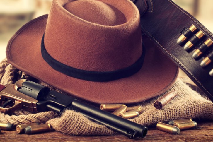 Cowboyhut liegt mit Lederpatronengürtel und Revolver auf einem Holztisch in rustikalem Licht.