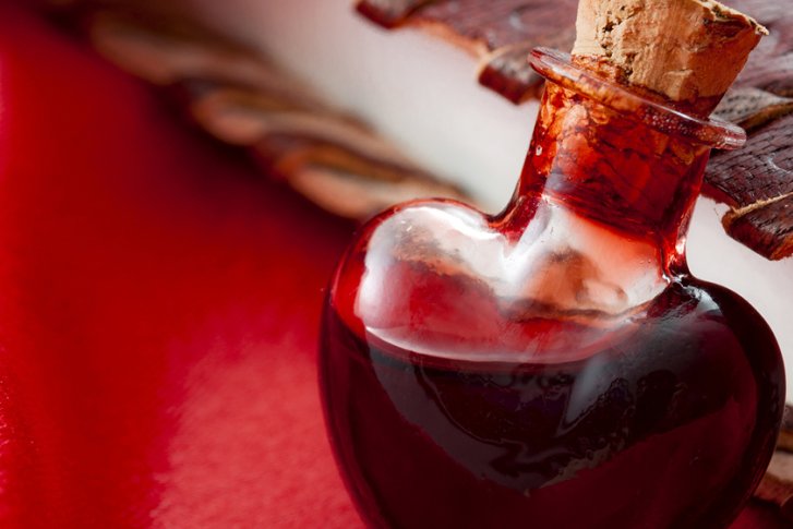 Herzförmige Glasflasche gefüllt mit roter Flüssigkeit liegt auf rotem Stoff neben einem Buch.