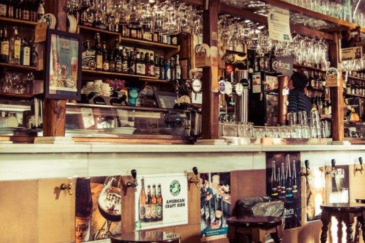 Bar mit Holztheke, Gläsern und Flaschen, umgeben von dekorativen Postern in gemütlicher Atmosphäre.