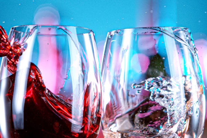 Zwei Gläser stoßen an, Flüssigkeiten spritzen, vor buntem, unscharfem Hintergrund.