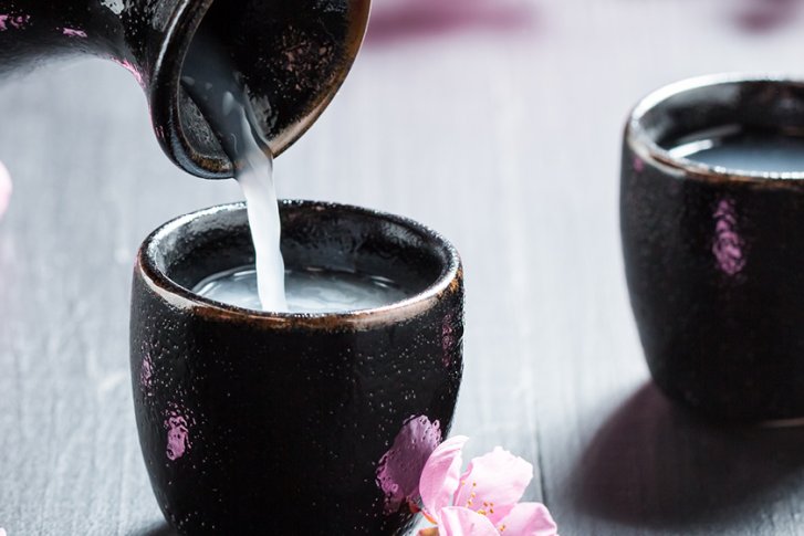 Zwei schwarze Tassen, Sake wird eingeschenkt, umgeben von rosa Blüten auf einem Holztisch.