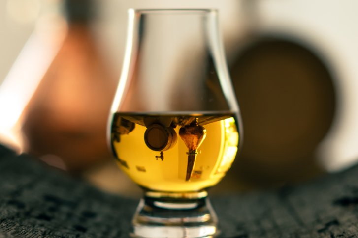 Ein halbvolles Whiskyglas spiegelt Fässer und Kupferkessel in einer rustikalen Umgebung wider.