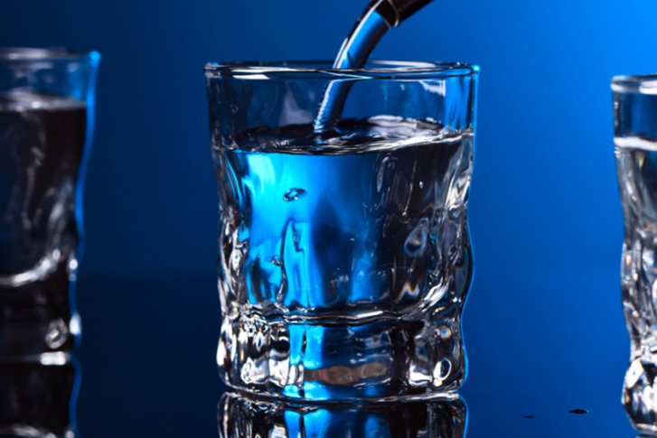 Drei Gläser stehen auf einer glänzenden Oberfläche, eines wird mit klarem Getränk gefüllt.