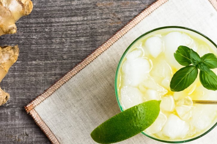 Ein Glas mit Eis und Limette steht auf einem Holztisch neben Ingwer und Basilikum.
