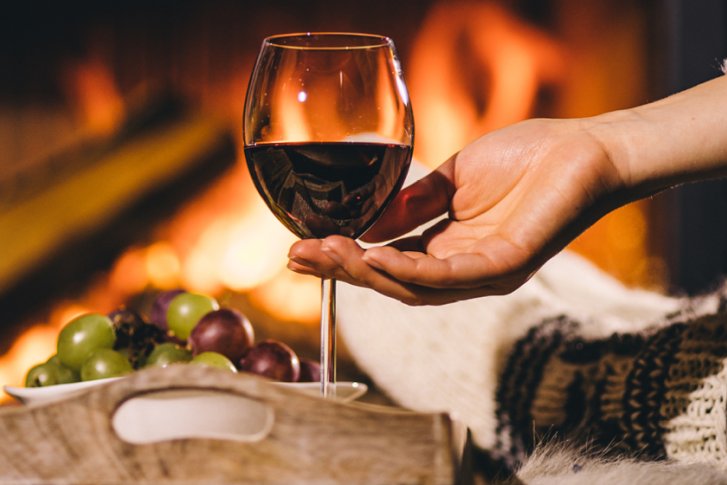 Weinglas wird gehalten, neben Tablett mit Trauben, vor gemütlichem Kaminfeuer.
