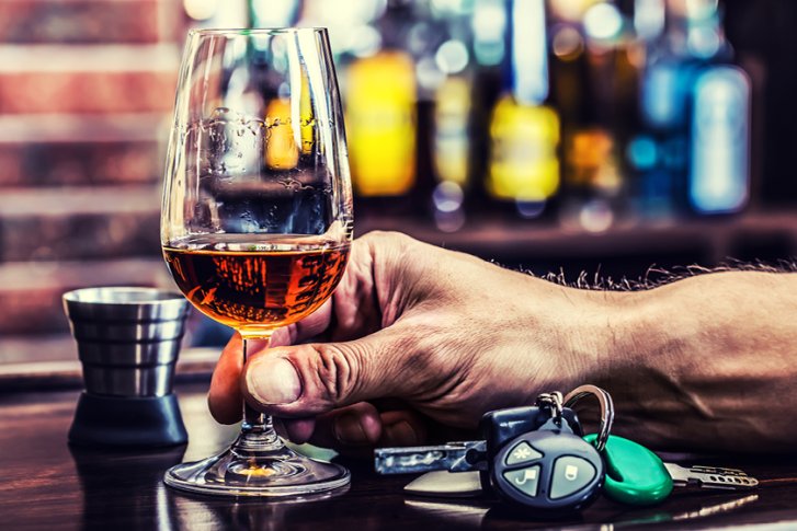 Ein Glas Whiskey in einer Hand, neben Autoschlüsseln, auf einer Baroberfläche.