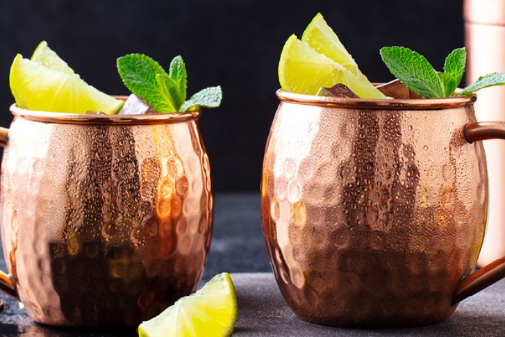 Zwei Kupferbecher, gefüllt mit Limetten und Minze, stehen auf einem Tisch mit schwarzem Hintergrund.