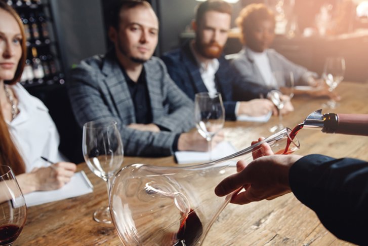 Karaffe wird mit Rotwein gefüllt, während vier Personen aufmerksam an einem Holztisch sitzen.
