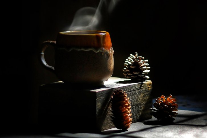 Eine dampfende Tasse steht auf einem Holzblock, umgeben von Tannenzapfen im Dunkeln.