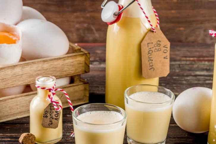 Flaschen mit Eierlikör und Gläser stehen neben einem Korb mit Eiern auf Holztisch.