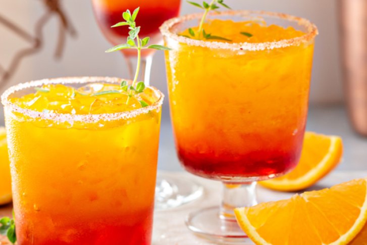 Zwei bunte Cocktails mit Eis und Kräuterdekoration, umgeben von Orangenscheiben, auf einem Tisch.