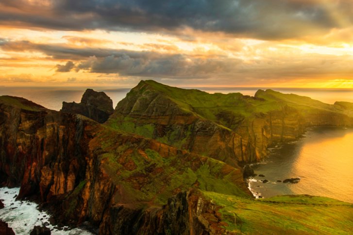 Felsige Küstenlandschaft bei Sonnenuntergang, grüne Hügel, ruhiges Meer und beleuchteter Himmel.