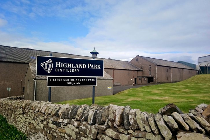Ein Schild mit "Highland Park Distillery" steht vor alten Lagergebäuden und einer Steinmauer.