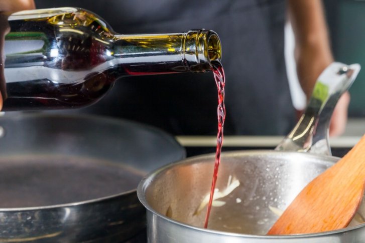 Hand gießt Rotwein aus Flasche in Topf, Holzlöffel daneben auf Herd.