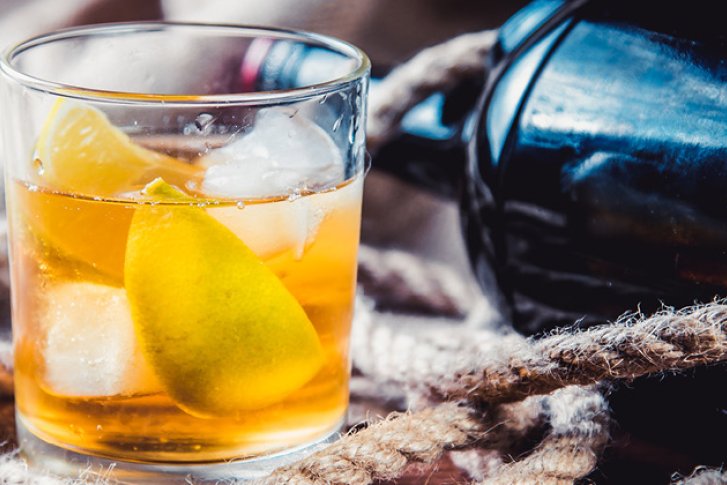 Glas mit Orangengetränk, Eiswürfeln und Zitronenscheibe steht auf Leinwand neben dunkler Flasche.
