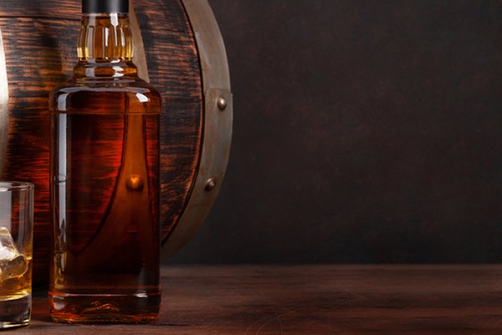 Glas Whisky mit Eiswürfeln steht vor Flasche und Holzfass auf Holztisch.