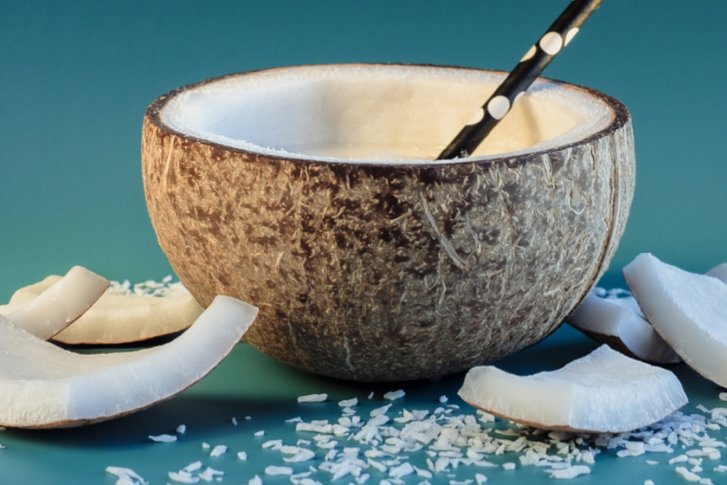 Kokosnussschale liegt mit Strohhalm und Kokosstücken auf blauem Untergrund.