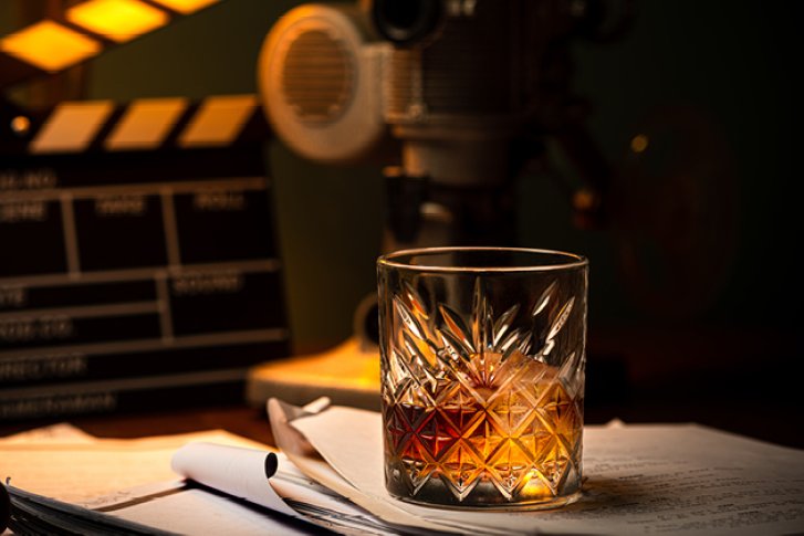 Ein Whiskyglas steht auf Papieren, umgeben von Filmkamera und Klappe, in gedimmter Beleuchtung.
