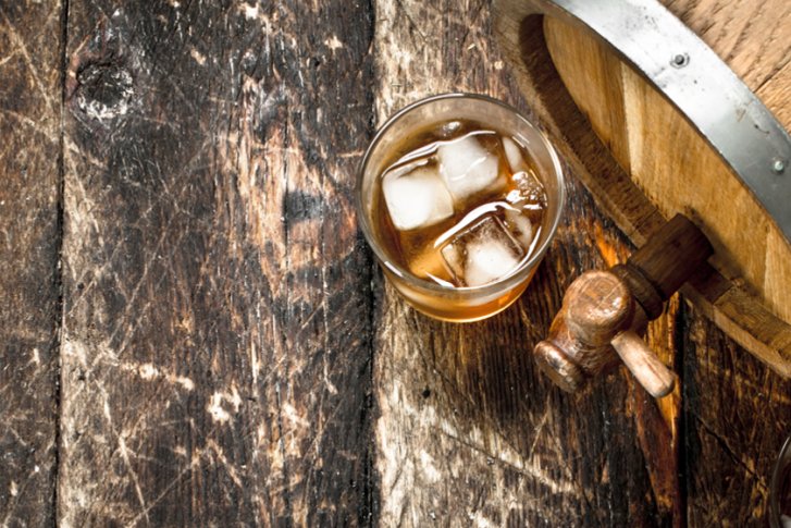 Ein Glas mit Eiswürfeln und Alkohol steht neben einem Holzfass, auf rustikalem Holztisch.