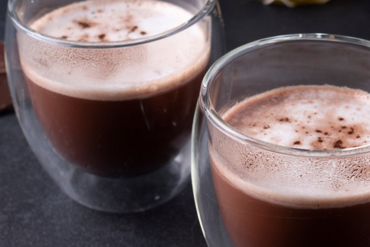 Zwei Gläser mit heißer Schokolade stehen auf dunklem Untergrund neben Schokoladenstücken.