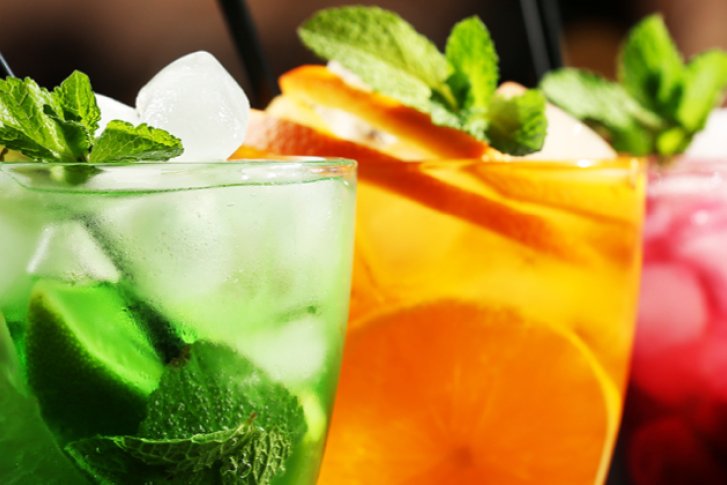 Drei bunte Cocktails mit Eiswürfeln stehen in einer Reihe vor unscharfem Hintergrund.