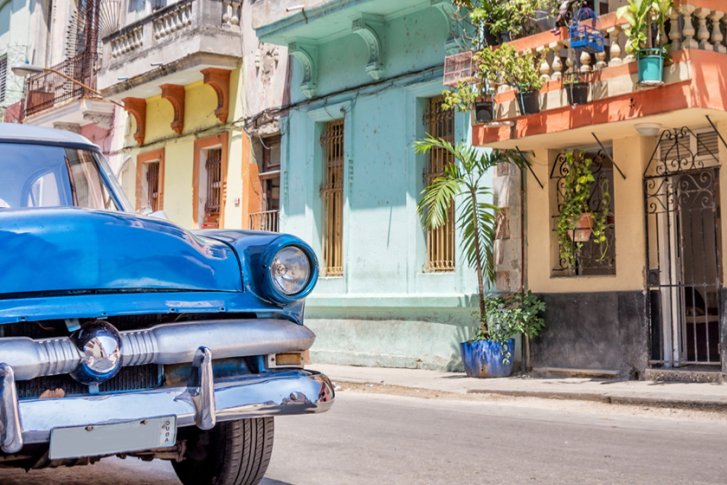 Blaues Vintage-Auto steht geparkt vor bunten Häusern in einer sonnigen Straßenkulisse.