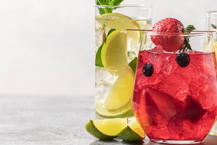 Drei bunte Cocktails, dekoriert mit Früchten, stehen auf einem grauen Tisch mit Zitrusfrüchten.