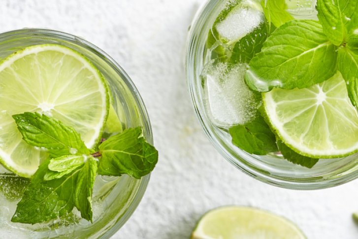 Zwei Gläser Mojito mit Limettenscheiben, Eis und Minze auf weißer, strukturierter Oberfläche.