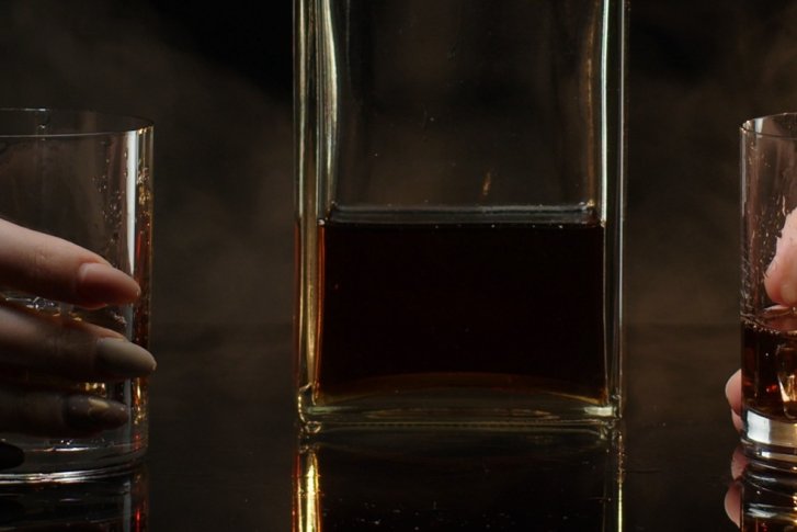 Zwei Hände halten Whiskygläser neben einer befüllten Flasche auf einem dunklen, reflektierenden Tisch.