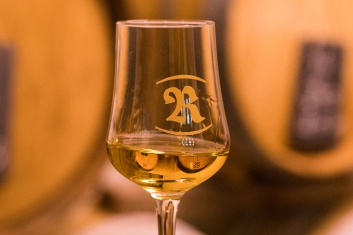 Ein Glas mit goldenem Getränk steht vor unscharfen hölzernen Fässern in warmem Licht.