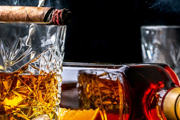 Glas Whisky mit Zigarre, daneben liegende Flasche und Orangenscheiben auf einem Tisch.