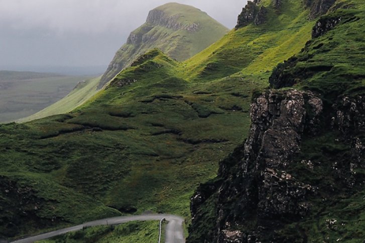 Grüne Hügel laufen über sanfte, kurvenreiche Straße, eingebettet in neblige, ruhige Landschaft.