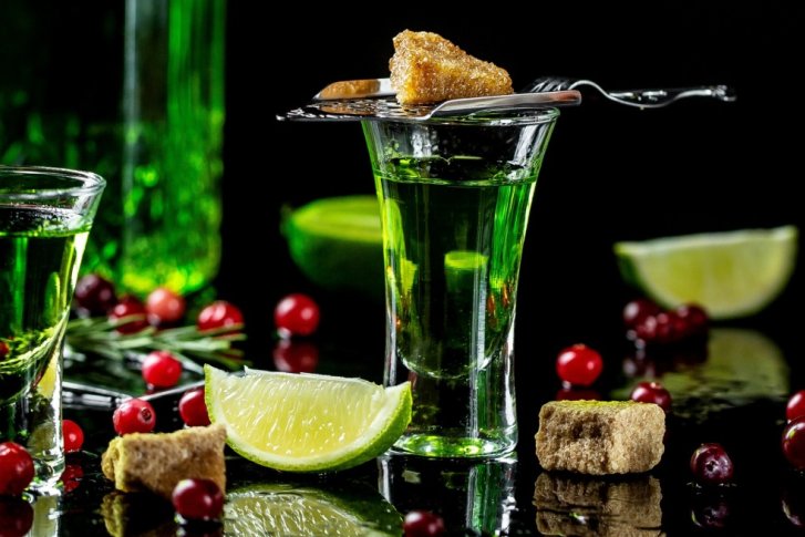 Grünes Getränk in einem Glas mit Zuckerwürfel daneben, umgeben von Limetten und Beeren.