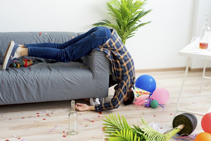 Person hängt kopfüber vom Sofa, umgeben von Konfetti, Ballons und leeren Flaschen.