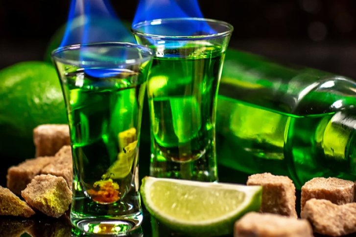 Zwei grüne Shots brennen, neben Limetten, Zuckerwürfeln und liegender Flasche auf schwarzer Oberfläche.