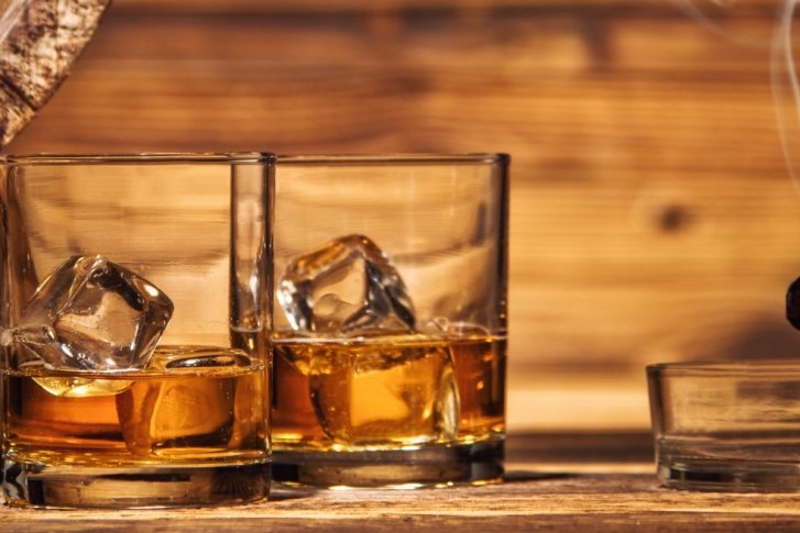Zwei Gläser mit Whiskey und Eis stehen neben einer rauchenden Zigarre auf Holztisch.