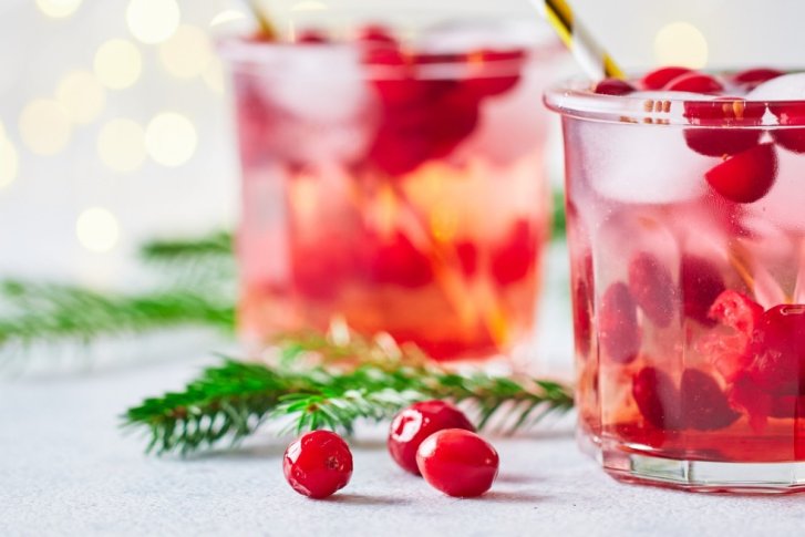 Zwei Gläser mit roten Getränken stehen auf einem Tisch, umgeben von Tannenzweigen und Lichtern.