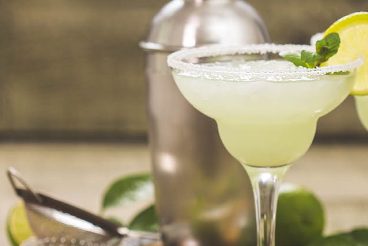 Zwei Margaritas mit Limettenscheiben stehen auf einem Tisch neben einem Cocktailshaker und Limettenblättern.