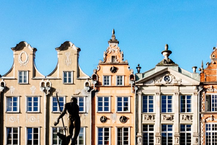 Reihen bunter, historischer Fassaden stehen nebeneinander. Eine Statue steht vorne, Himmel dahinter.