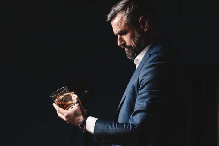 Mann betrachtet Drink in einem Glas, trägt Anzug, vor dunklem Hintergrund, Seitenprofil.