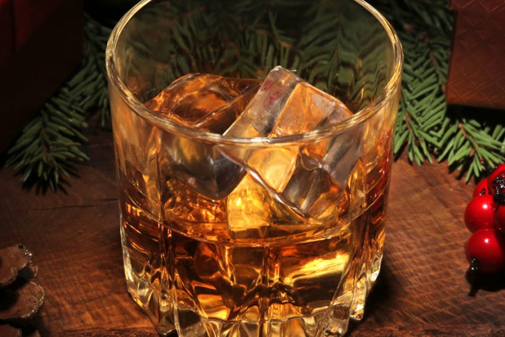 Whiskeyglas mit Eiswürfeln steht auf Holztisch, dekoriert mit Tannenzweigen und roten Beeren.