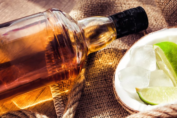 Flasche liegt neben halber Kokosnuss mit Eis und Limetten auf Jutestoff und Holzuntergrund.