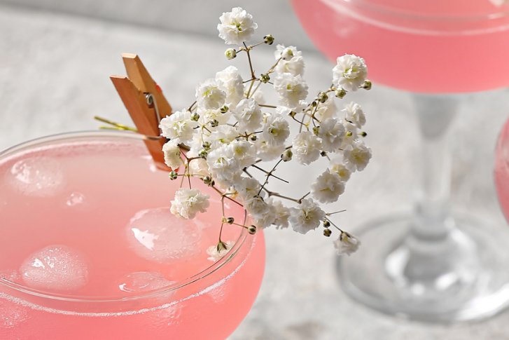 Rosa Cocktails stehen dekoriert mit Blumen und Eiswürfeln auf einer grauen Tischoberfläche.
