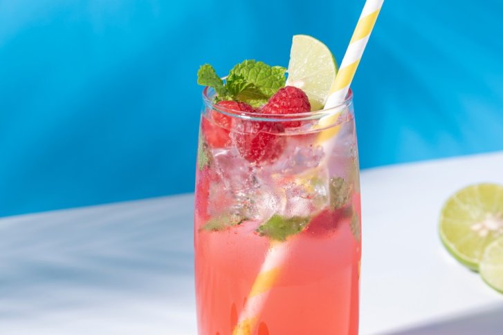 Rosa Cocktail mit Himbeeren, Minze und Strohhalm steht auf einem Tisch, blaue Hintergrund.