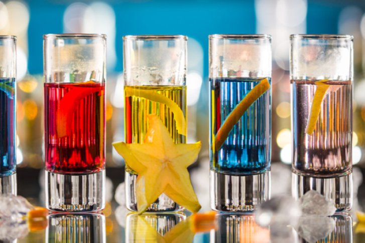 Bunte Cocktails in Gläsern, dekoriert mit Obstscheiben, stehen auf spiegelnder Oberfläche, Hintergrund verschwommen.