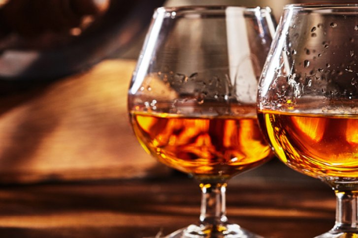 Zwei gefüllte Cognacgläser stehen auf einem Holztisch neben einem unscharfen Fass im Hintergrund.